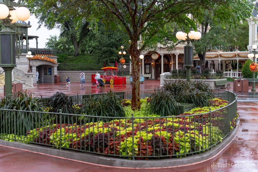 A landscaped area with colorful plants and flowers, surrounded by a green iron fence, in a park-like setting. A bright red food cart is visible nearby, with a few people walking under a cloudy sky. The area is wet, suggesting recent rain.