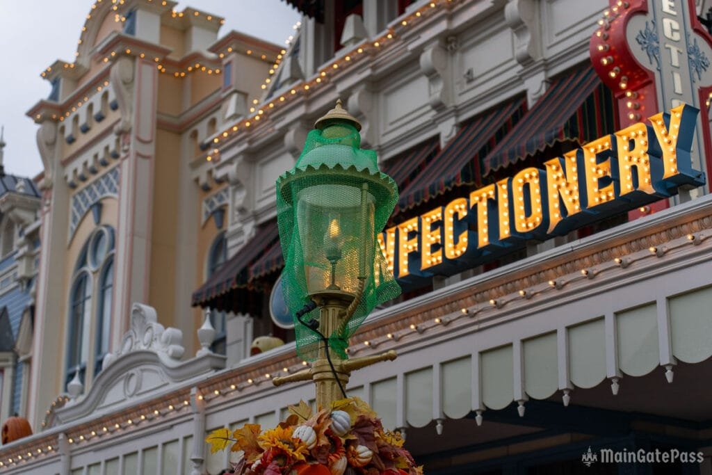 A decorative street lamp draped in green mesh stands in the foreground, with a building featuring a brightly lit sign that reads "INFECTIONERY" in the background, adorned with autumn decorations and festive lights.