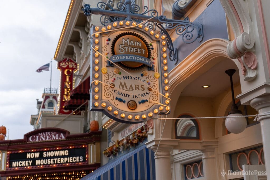 Sign for "Main Street Confectionery," advertising it as the home of Mars candy treats, with decorative lights and colorful graphics. In the background, a marquee for "Main St Cinema" showing "Now Showing Mickey Mousepieces" is visible.