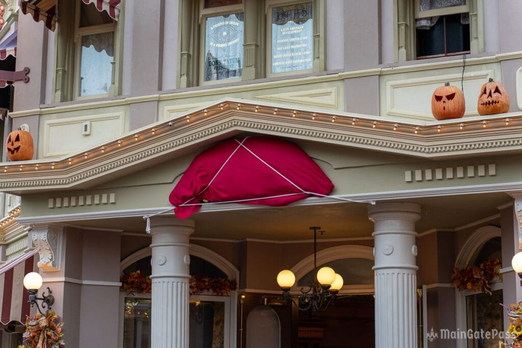 A red cloth is draped over a decorative feature above the entrance of a building, with two carved pumpkins placed on the roof above. The building has columns and is adorned with strings of lights and autumn-themed decorations.