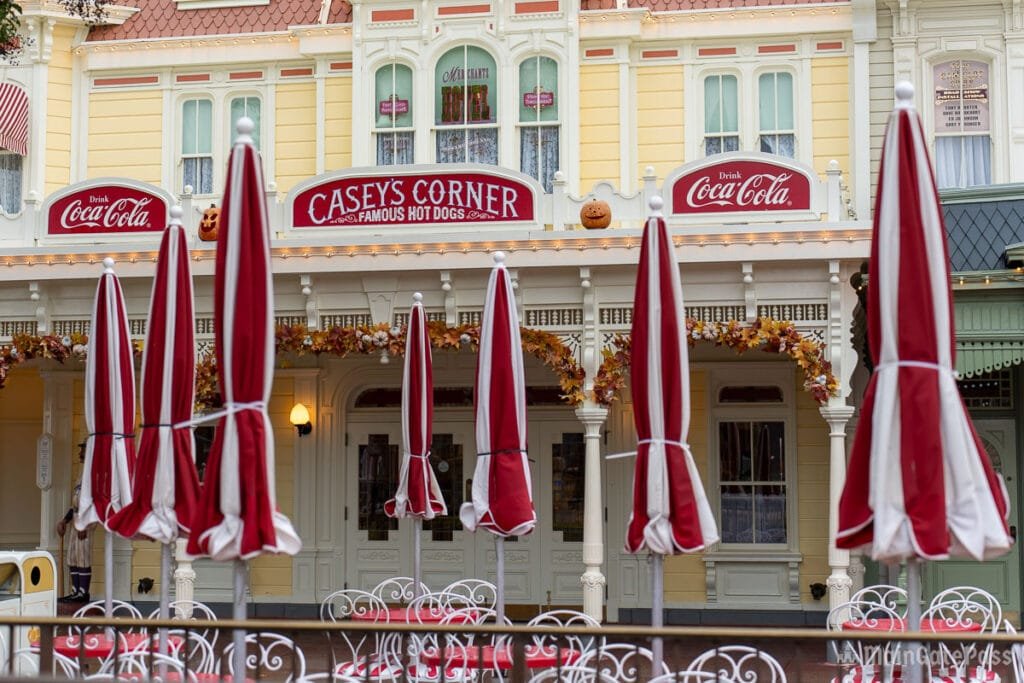 The exterior of Casey's Corner, a hot dog restaurant, featuring red and white striped umbrellas, decorative autumn garlands, and a Coca-Cola sign.