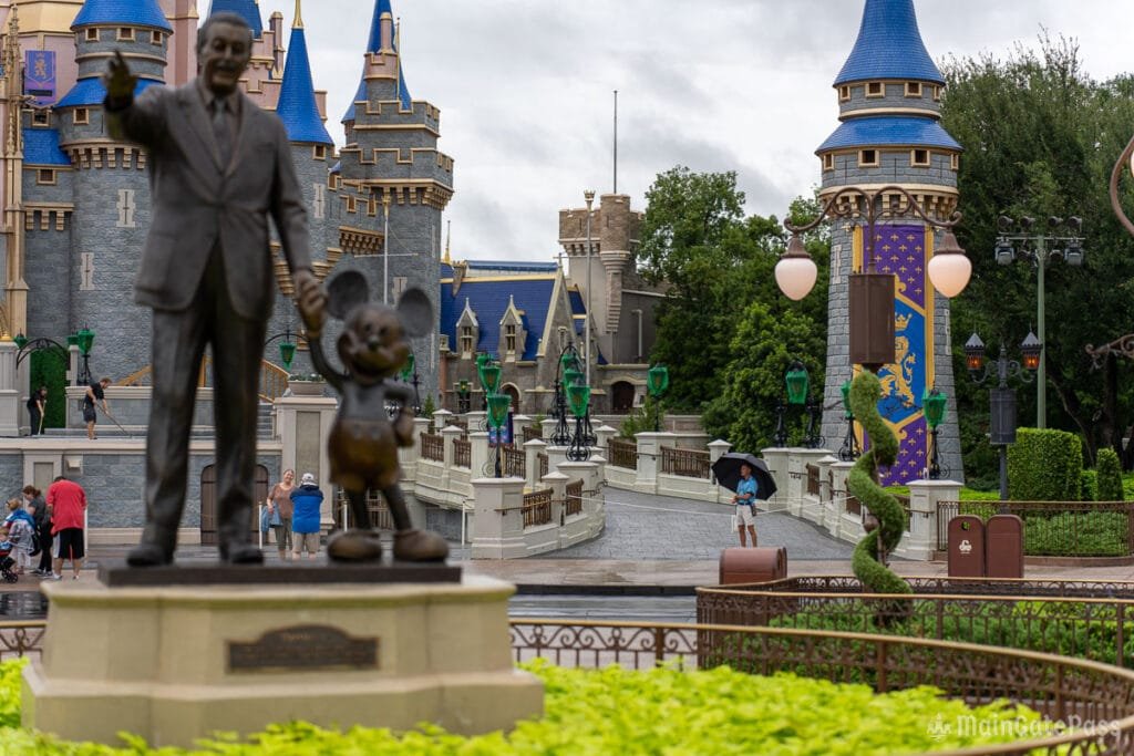 A bronze statue of Walt Disney and Mickey Mouse stands in the foreground, with a castle featuring blue spires in the background. Several people are walking in the area, including a child holding an umbrella, while lush green plants frame the scene.