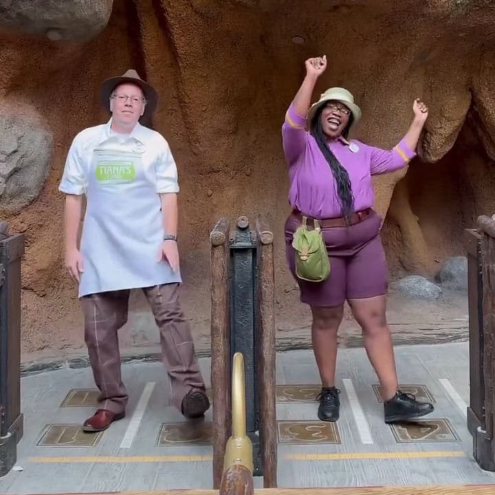 Two individuals are standing in front of a rocky background, posing in a playful manner. One is wearing an apron and a hat, with a serious expression, while the other is wearing a purple shirt and shorts, smiling and raising their arms in excitement.