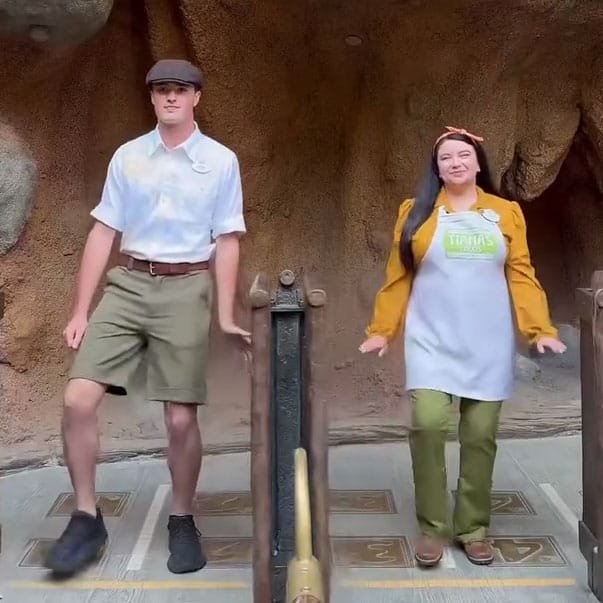 Two individuals are dancing in front of a textured rock background. The male, dressed in a white shirt and green shorts with a cap, stands on the left. The female, wearing an orange shirt over an apron and green pants, stands on the right, smiling at the camera.