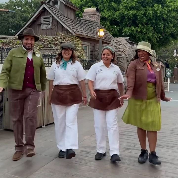 Four individuals dressed in themed costumes are happily walking and engaging with each other in an outdoor setting, with a rustic building and trees in the background.