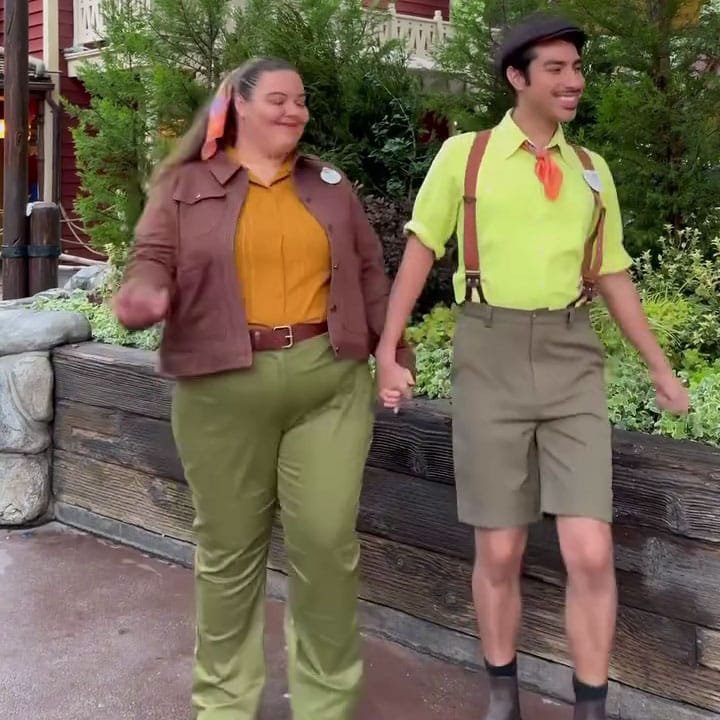 Two people are walking hand in hand outdoors. One person is wearing a brown jacket, orange shirt, and green pants, while the other is dressed in a bright yellow shirt, suspenders, and green shorts. They appear to be smiling and enjoying each other's company, with greenery and wooden structures in the background.