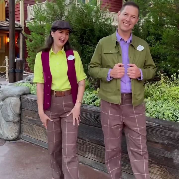 Two people pose for a photo outdoors, both wearing colorful outfits. The woman on the left sports a bright lime green shirt with a maroon vest and plaid pants, while the man on the right wears a green jacket over a purple shirt, paired with similar plaid pants. There are trees and a wooden structure in the background.