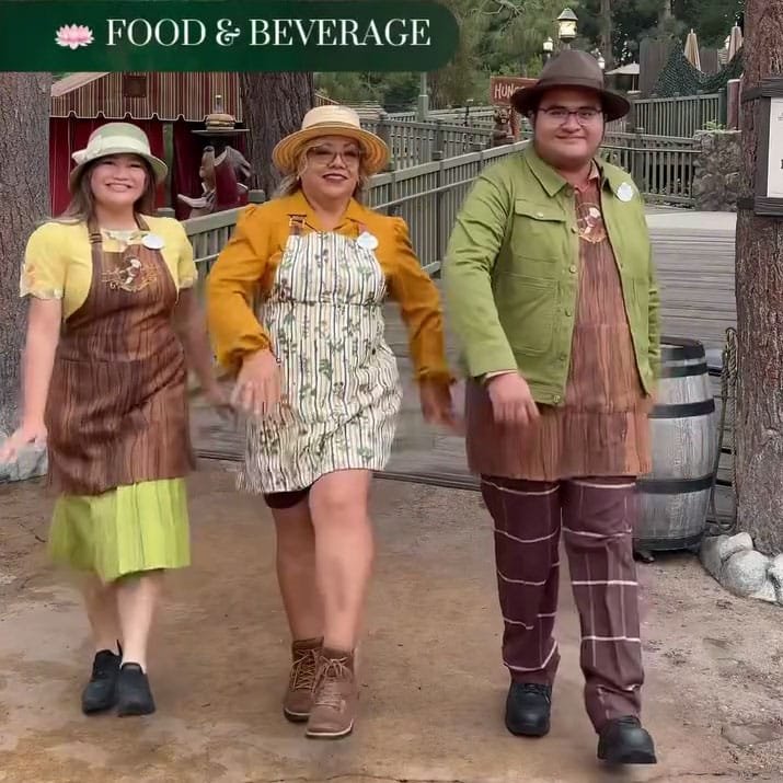 Three individuals are walking together outdoors, dressed in coordinating, rustic-style outfits. The woman in the center wears a patterned apron dress with long sleeves and glasses, flanked by two others: one in a brown overall and a yellow blouse, and the other in a green jacket with patterned pants. The background features trees and outdoor decor.
