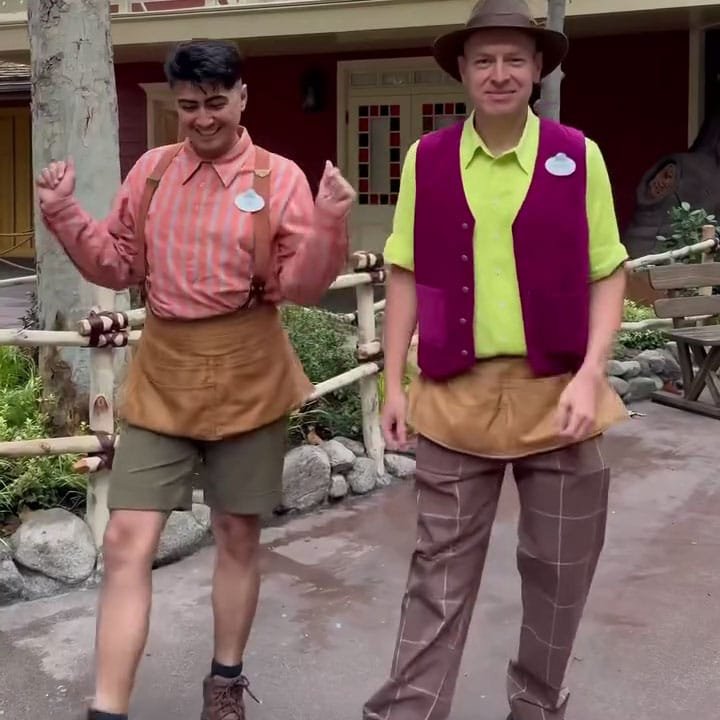 Two men dressed in colorful costumes are dancing outdoors. One wears a striped pink shirt, shorts, and a brown apron, while the other wears a bright yellow shirt, a purple vest, and plaid pants. The background features a rustic environment with trees and decorative elements.