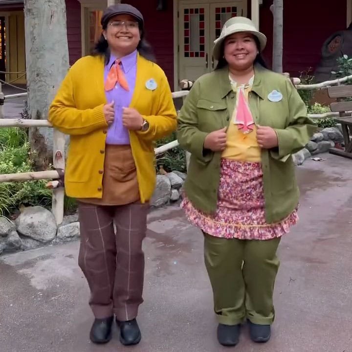 Two women stand smiling in a theme park setting, dressed in colorful outfits. One wears a yellow cardigan over a purple top, brown pants, and black shoes, while the other is in a green jacket over a yellow top, floral skirt, and green pants. Both have accessories like scarves and hats.