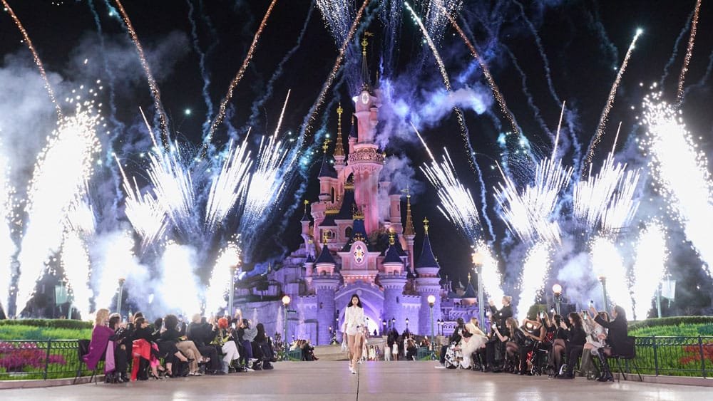 A beautiful night view of a castle with fireworks illuminating the sky, while an audience sits and watches the display in front of it.