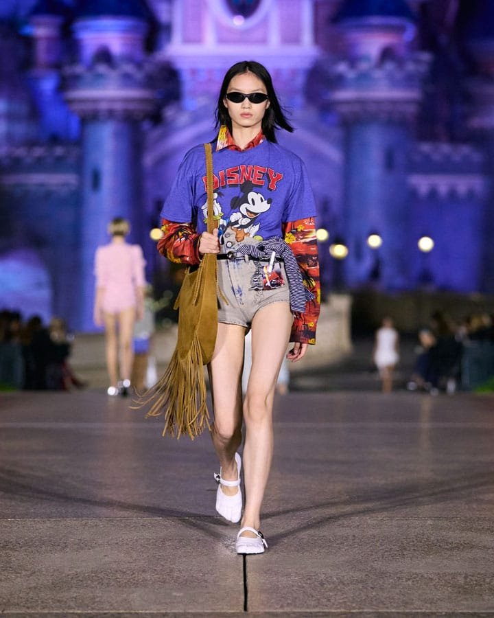 A model walks down a runway wearing a colorful oversized Disney-themed t-shirt, denim shorts, and sunglasses, with a fringed brown bag slung over her shoulder. The backdrop features a vibrant castle illuminated, enhancing the fashion show atmosphere.