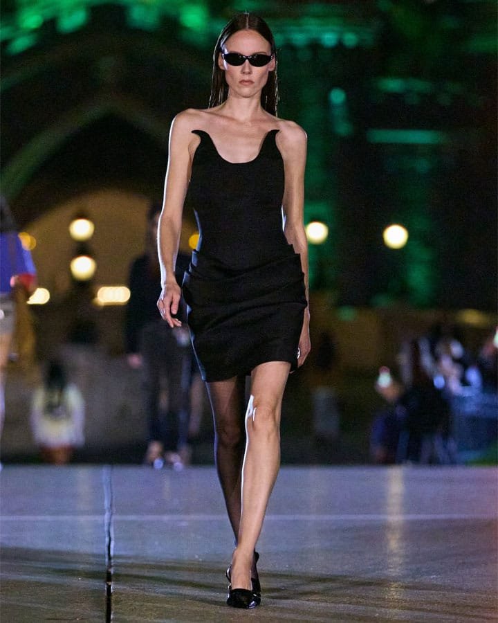 A model walks down a runway wearing a sleek black dress and sunglasses, with a nighttime atmosphere featuring blurred lights in the background.