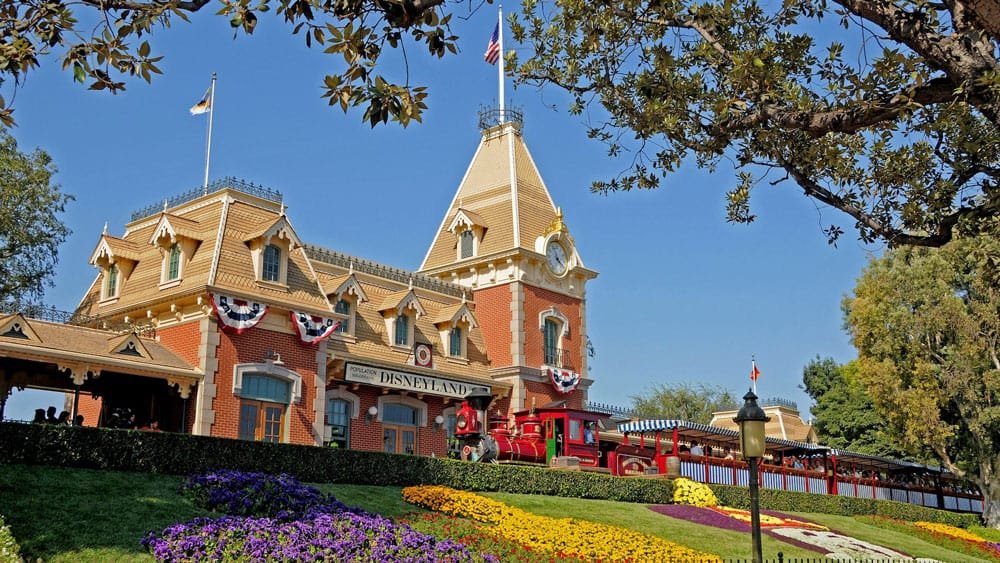 A picturesque view of the Disneyland entrance featuring a large, ornate brick train station with a peaked roof, flags flying, and colorful flower beds in the foreground. A red vintage train is parked at the station, under a clear blue sky.