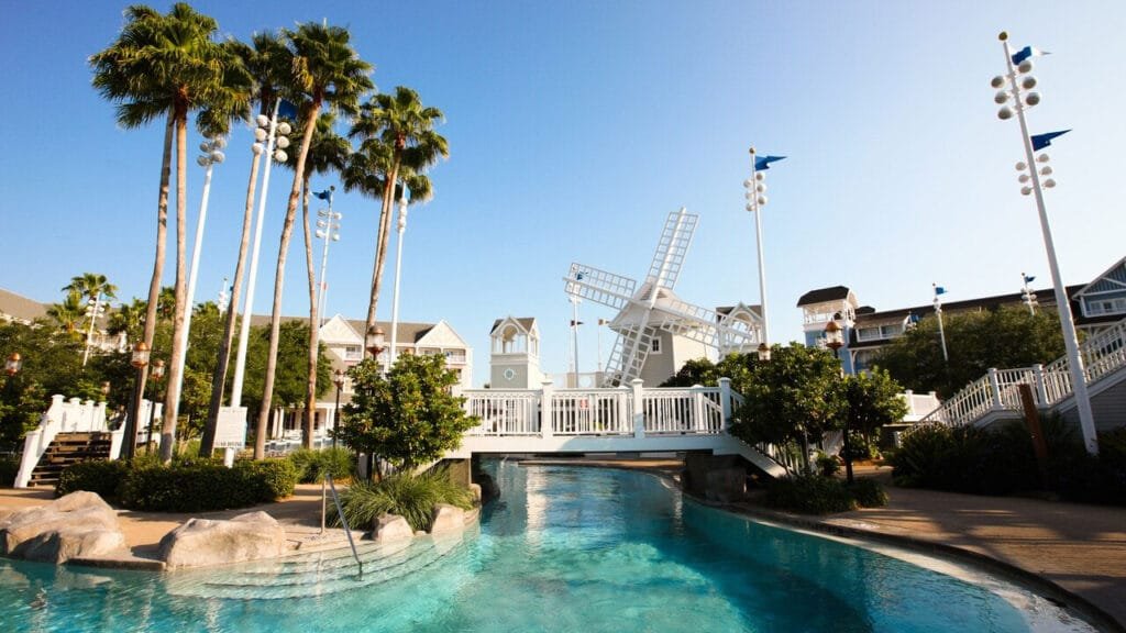 A scenic view of a resort area featuring tall palm trees, a white windmill, and a blue waterway with a wooden bridge. Surrounding buildings are designed in a coastal style under a clear blue sky.