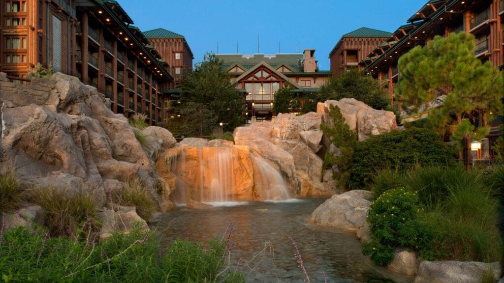 A peaceful outdoor scene featuring a cascading waterfall flowing over rocks, surrounded by lush greenery, with a rustic lodge building in the background and a twilight sky.
