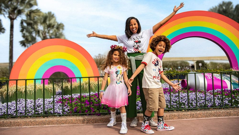 A woman and two children pose joyfully in front of colorful rainbow arches, surrounded by flowers at a theme park. The woman is wearing a Minnie Mouse t-shirt, while the children are dressed in playful outfits.
