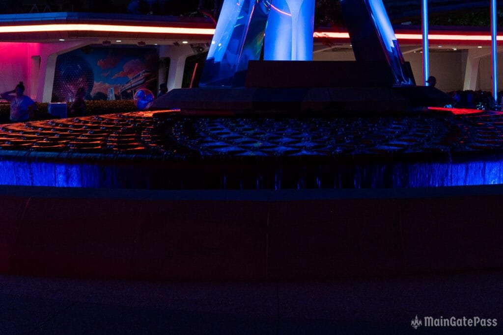 A brightly lit water feature with circular patterns, illuminated in blue and orange tones, set against a dark background with abstract artworks and structures partially visible.