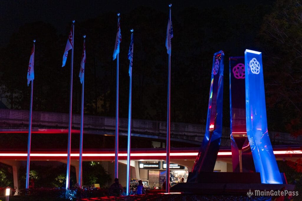 A nighttime scene featuring tall, illuminated flagpoles with colorful flags and a modern, abstract monument with blue and purple lighting, set against a dark background. The area appears to be a public space with some people visible.