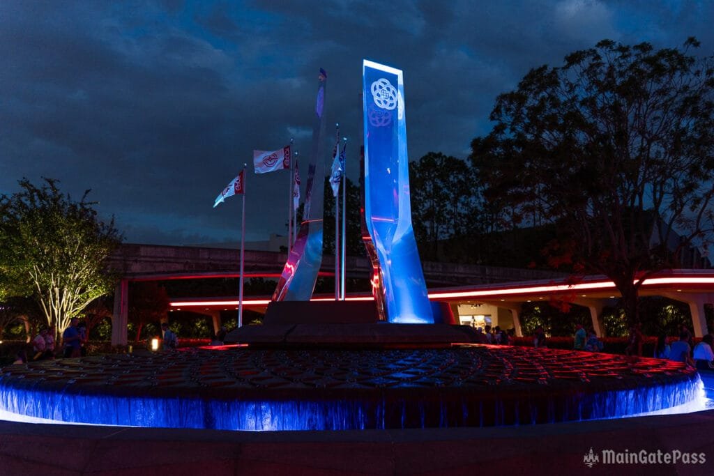 A nighttime view of a park area featuring illuminated blue and red fountains with tall, transparent pillars in the background and flags waving in the breeze. Trees and people can be seen around the fountain area.