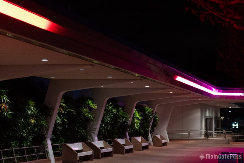 A dimly lit modern architectural space featuring a smooth, curved structure with a purple and red light strip along the top, surrounded by lush green plants and a few empty benches.