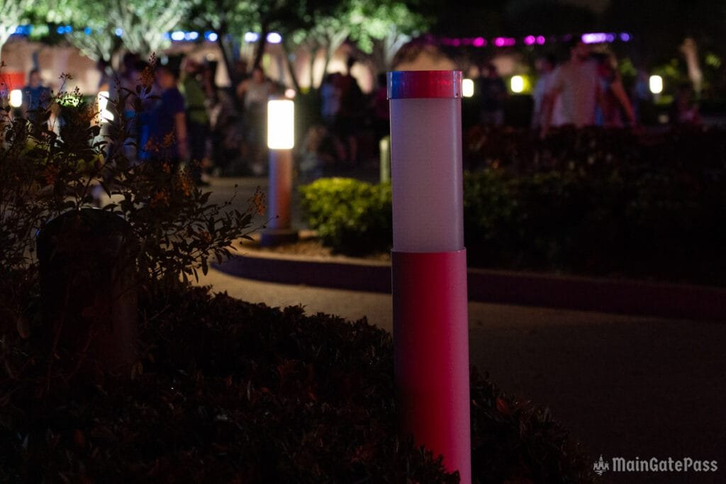 A dimly lit scene featuring a decorative light pole with red and white segments, surrounded by leaves and shrubs. In the background, groups of people are visible, with colorful lights illuminating the area.