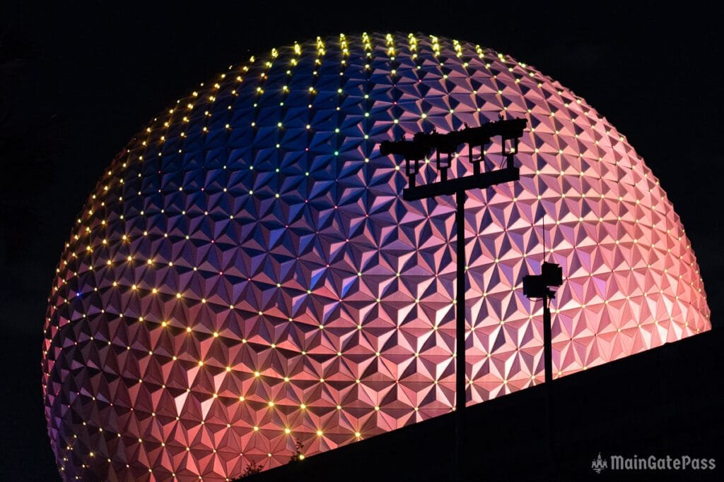 A brightly lit, multicolored geometric dome structure at night, with a silhouette of a tall pole and lights in the foreground.