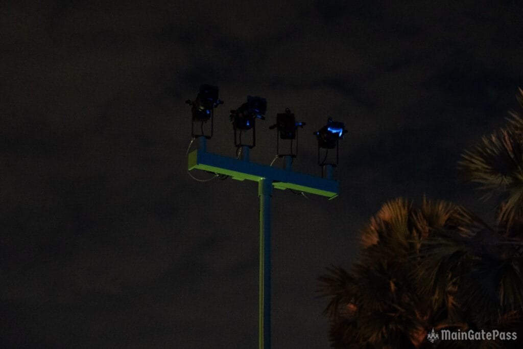 Four spotlights mounted on a tall pole, illuminated against a dark sky with a few clouds, and silhouette of palm leaves in the foreground.