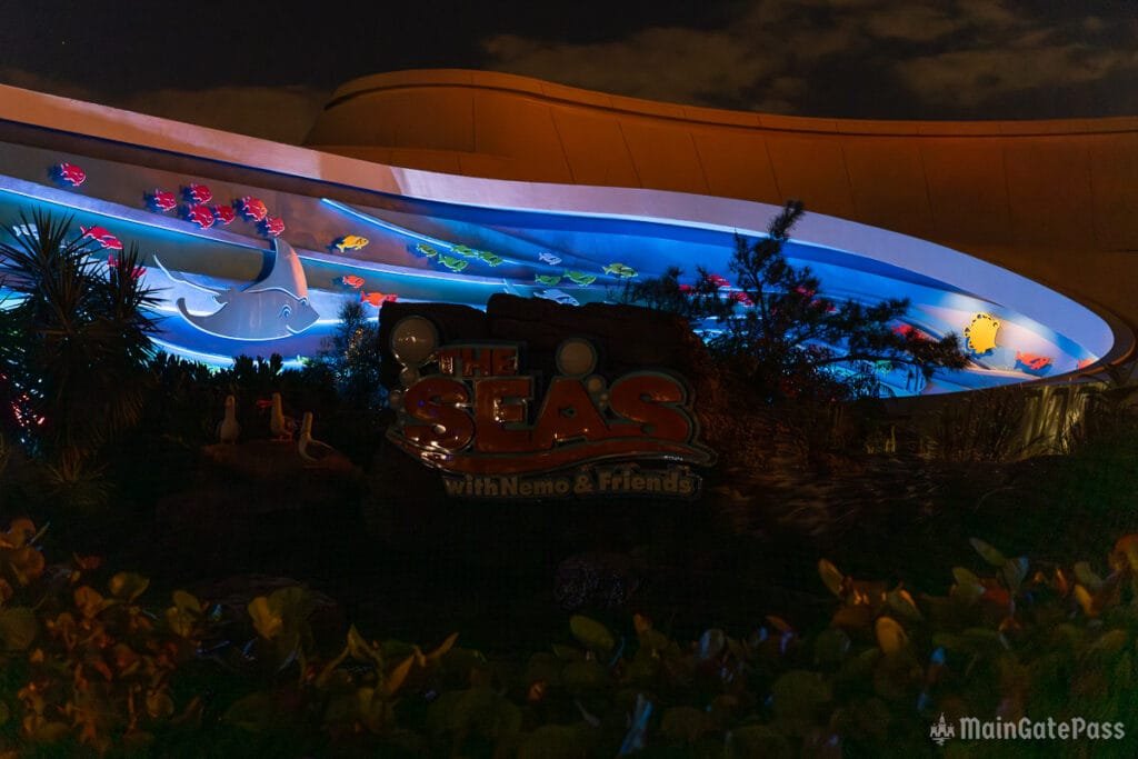 Colorful animated fish swimming under a lit archway showcasing the attraction sign "The Seas with Nemo & Friends" in a nighttime setting surrounded by greenery.
