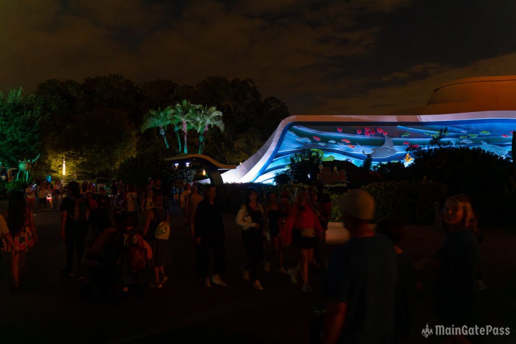 A nighttime scene at a theme park featuring a decorative illuminated aquarium mural with colorful fish, surrounded by palm trees and groups of people walking nearby.