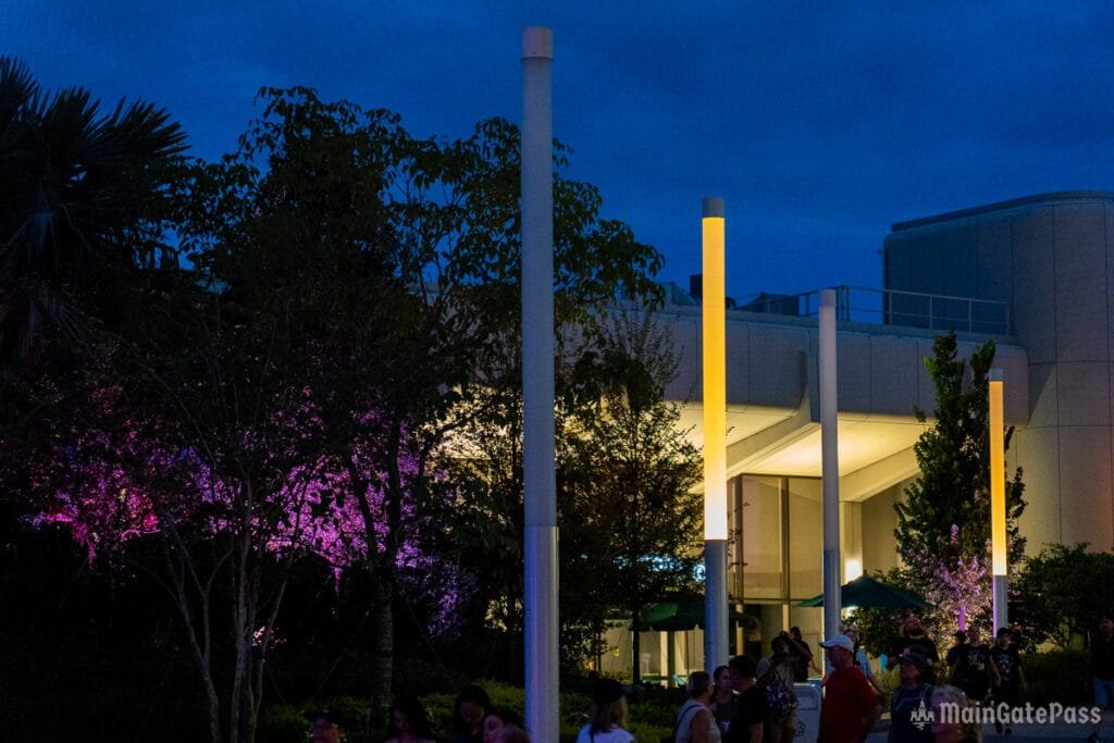 A nighttime scene featuring illuminated trees with purple and yellow lighting, alongside modern architectural elements and people walking in an outdoor area. The sky is darkening with twilight.