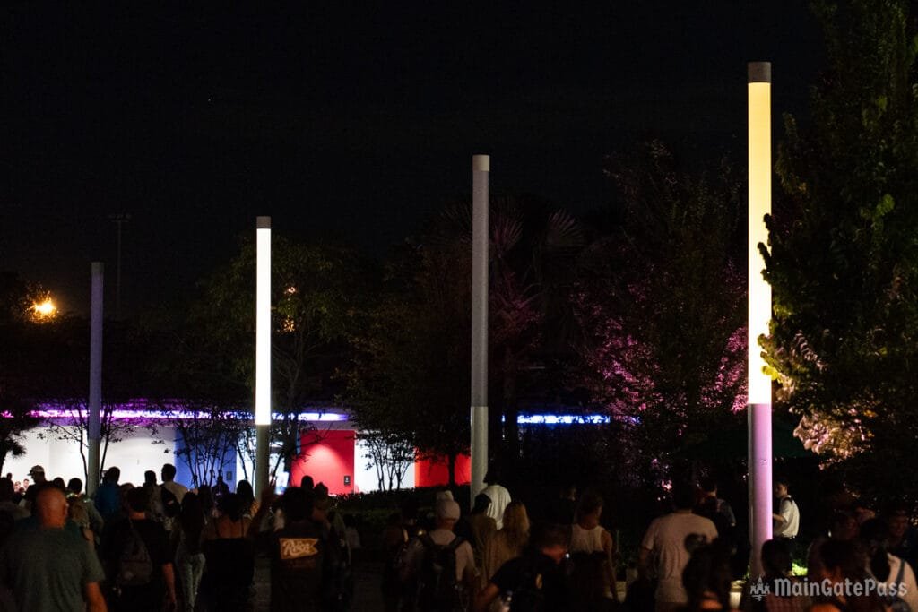 A crowd of people walks through a dimly lit area at night, surrounded by tall illuminated poles and colorful lighted structures in the background.