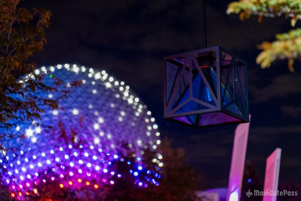 A decorative lantern hangs in the foreground, with a colorful, illuminated dome structure blurred in the background under a night sky.