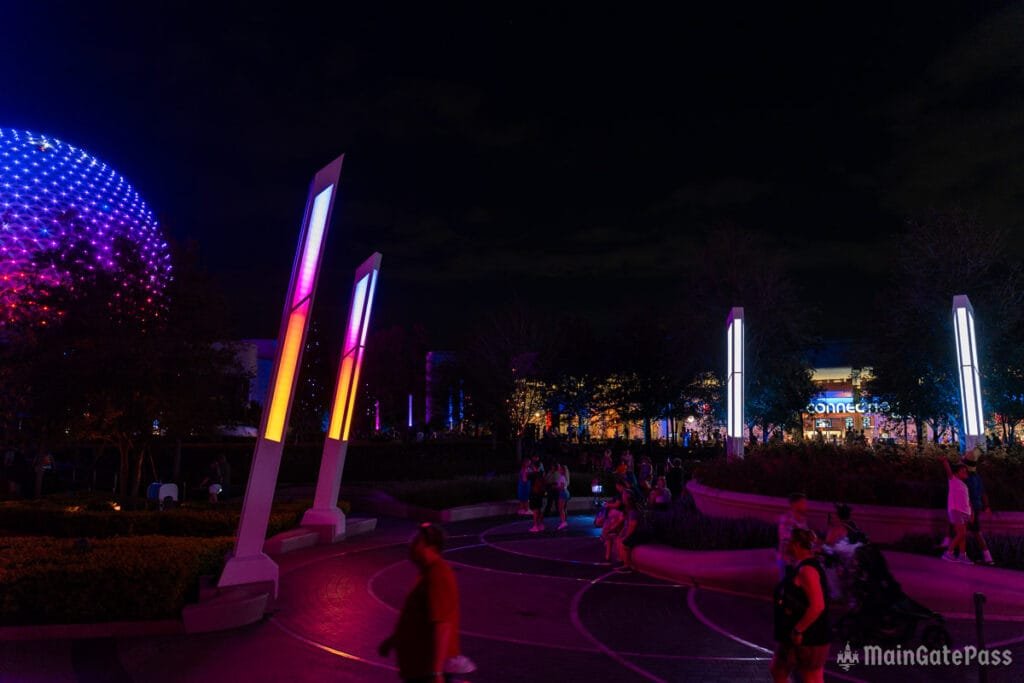 A nighttime scene at a theme park featuring colorful illuminated structures and a large, glowing sphere in the background. People are walking along a curved pathway, with trees and the facade of a building visible behind them.