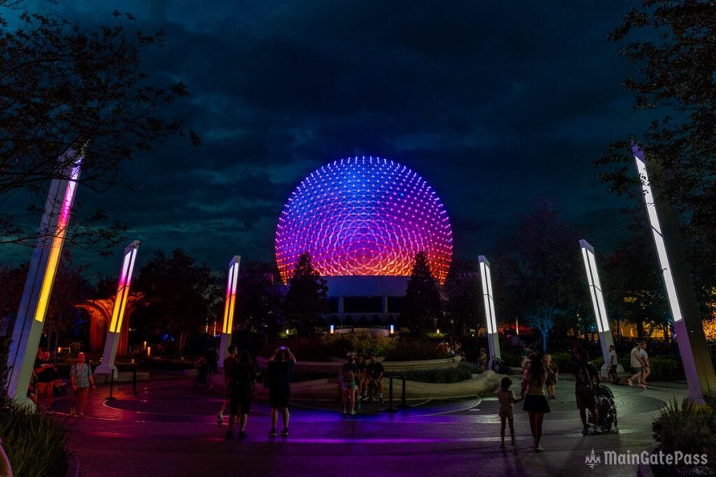 A colorful, illuminated sphere at night, surrounded by people walking and standing in a park-like setting, with trees and modern light structures.