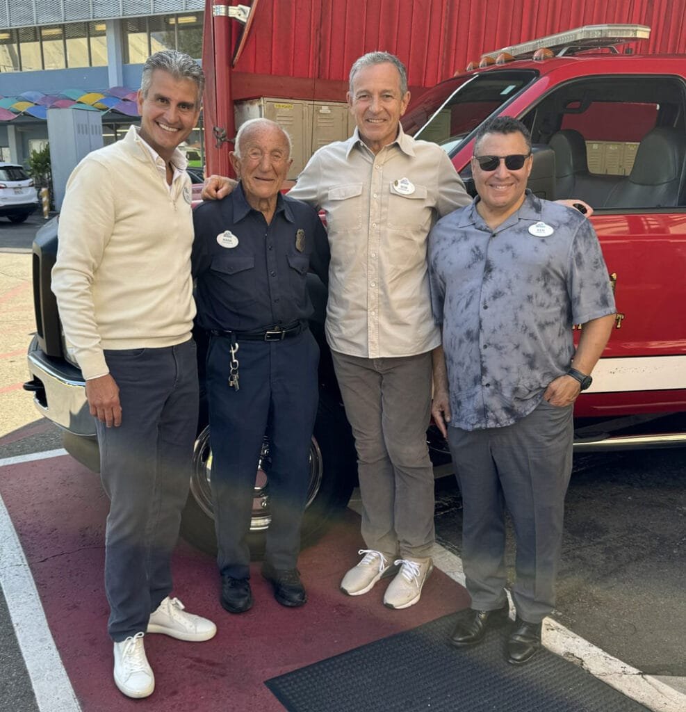 Four men pose together in front of a red fire truck. They smile and stand closely, with the truck and a colorful building in the background.