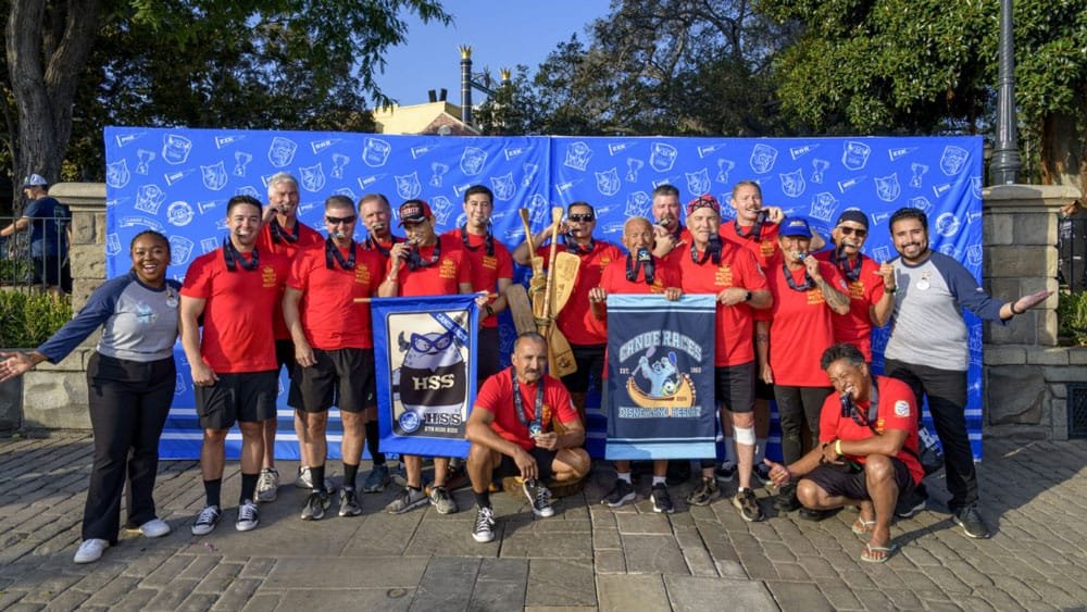 A group of people wearing red shirts and medals poses in front of a blue backdrop, holding a banner. They appear to be celebrating a team achievement, with a mix of genders and ages among them.