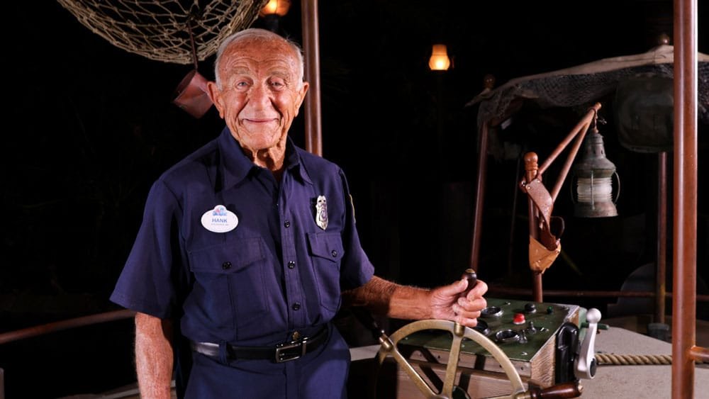 An elderly man in a navy blue uniform with a name tag smiles while standing next to a ship's wheel and control panel, with nautical decor in the background.
