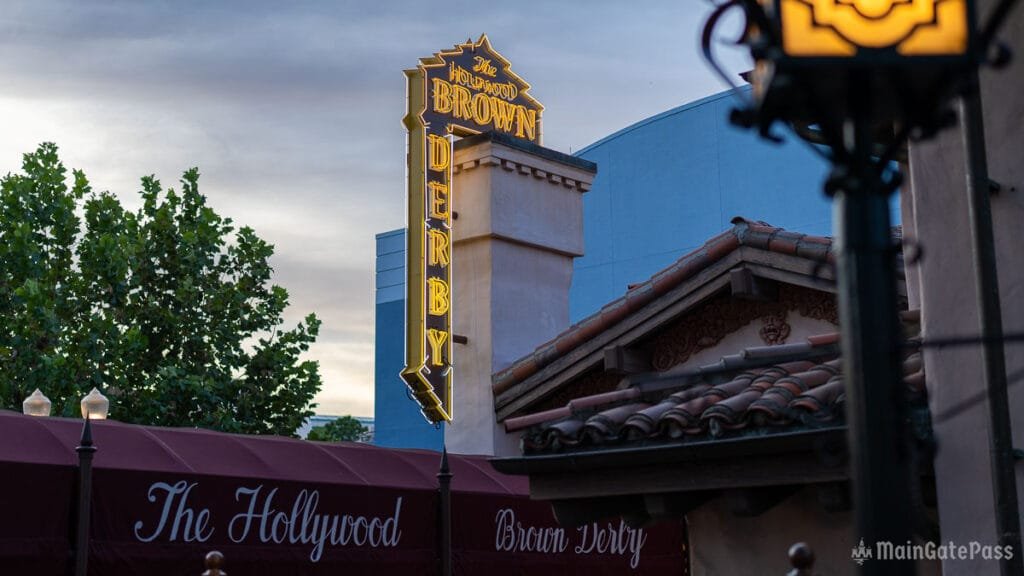 A vintage-style neon sign reading "The Hollywood Brown Derby" is illuminated against a dusky sky, with tree foliage in the foreground and a part of a building roof visible in the bottom.