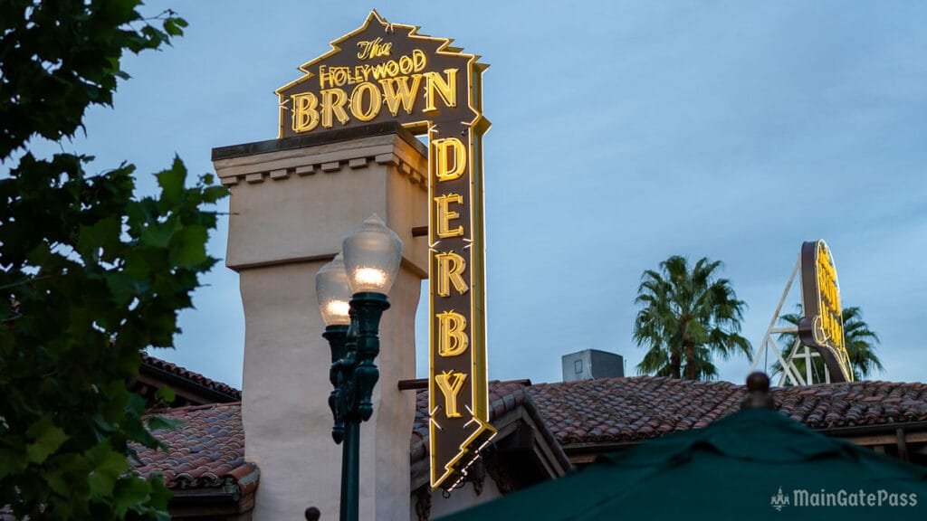 Sign for "The Hollywood Brown Derby" illuminated with neon lights, situated above a building, with palm trees and a streetlamp visible in the foreground against an evening sky.