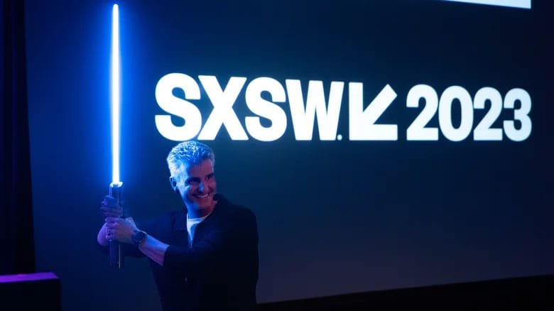 A person holding a glowing blue lightsaber in front of a large screen displaying the text "SXSW 2023."