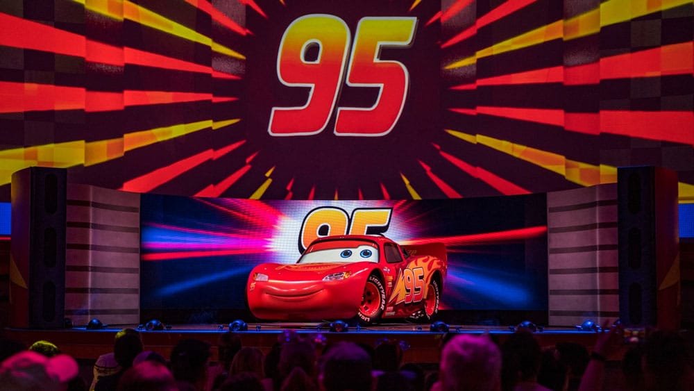 A brightly colored animated car with the number "95" on its side is on stage in front of a vibrant backdrop with graphic elements radiating outward. The audience is partially visible in the foreground.