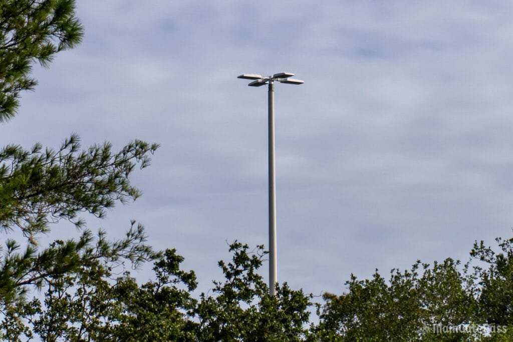 A tall, modern streetlight is seen rising above a line of trees under a cloudy sky.