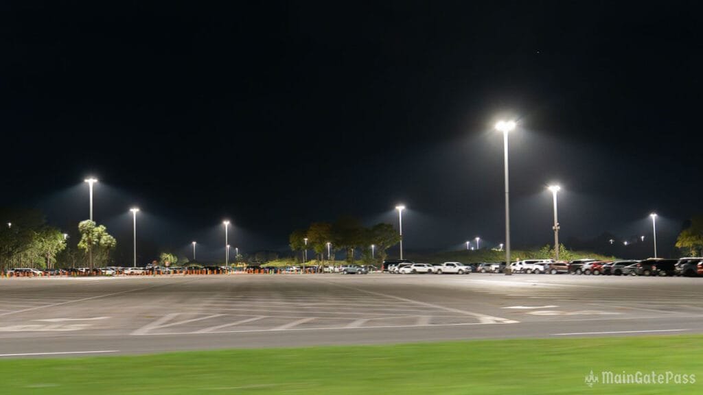 A dimly lit parking lot at night, featuring multiple streetlights illuminating parked cars and tree silhouettes in the background.