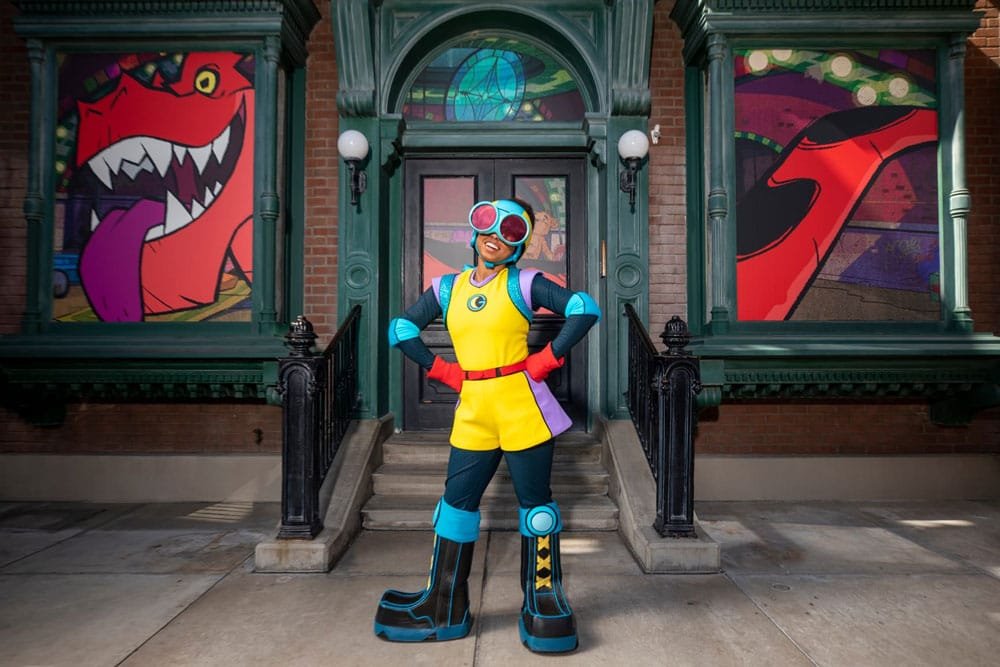 A person in a colorful superhero outfit poses confidently in front of a brick building with cartoonish monster murals on the windows.