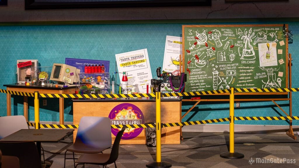 A table with various science experiment props and displays related to Muppet Labs, featuring colorful beakers, a chalkboard with drawings of experiments like "volcano" and "spaghetti bowling," and safety barriers with yellow and black stripes.