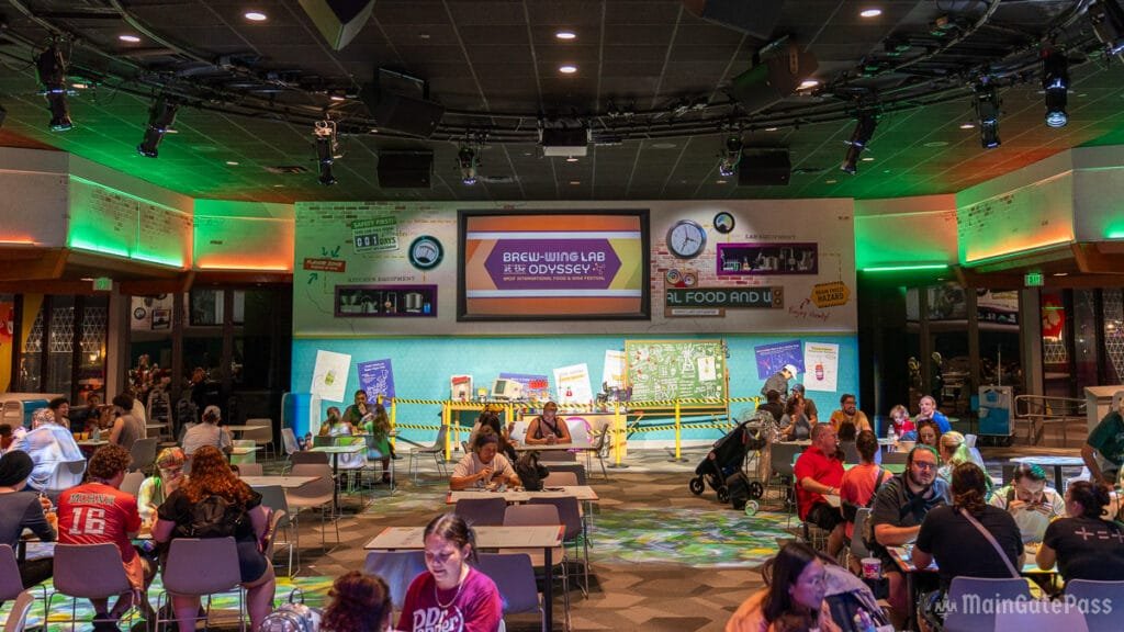 A vibrant indoor space labeled "Brew-Wing Lab at the Odyssey," featuring tables occupied by families and groups. The walls are decorated with colorful graphics and signs related to food and science. A large screen displays event information, and a mix of ambient lighting creates a lively atmosphere.