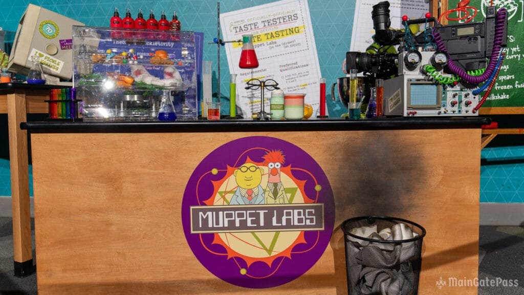 A tabletop display with colorful laboratory equipment and a trash bin, featuring a "Muppet Labs" logo with cartoon characters, surrounded by various bottles and scientific instruments against a decorative background.