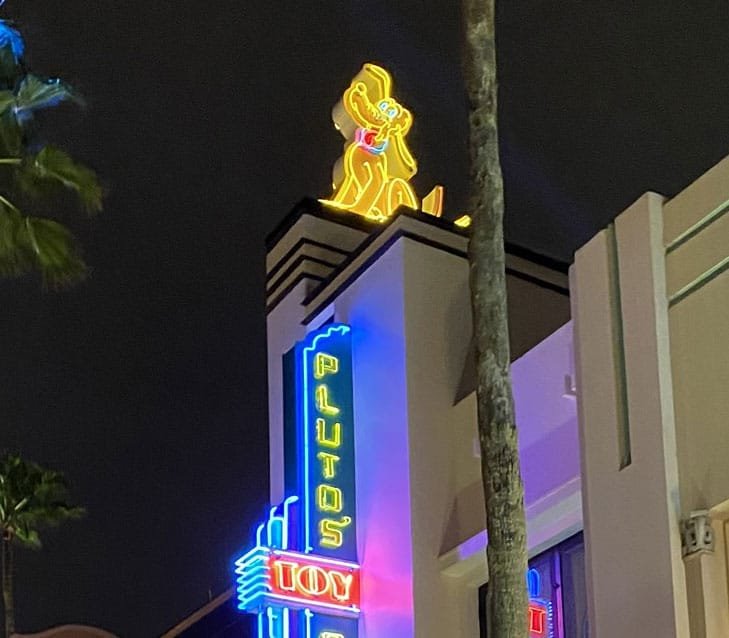 A brightly lit neon sign features a cartoon dog on top with the text "PLUTO'S TOY" in colorful lights, set against a night sky.