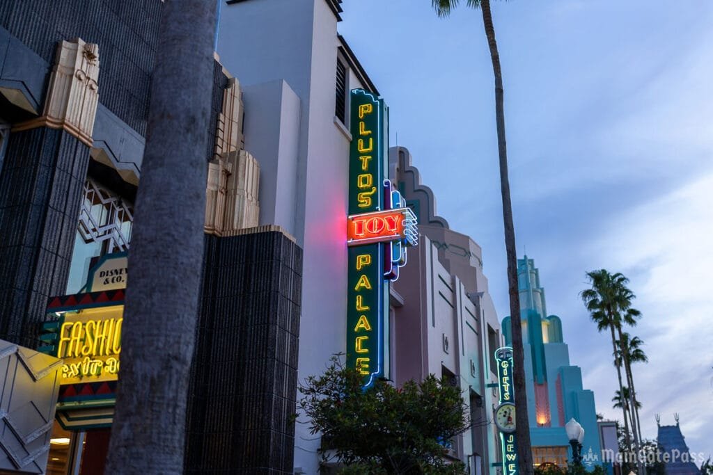 A brightly lit neon sign for "Pluto's Toy Palace" is visible above a storefront, with palm trees and Art Deco-style architecture in the background during dusk.